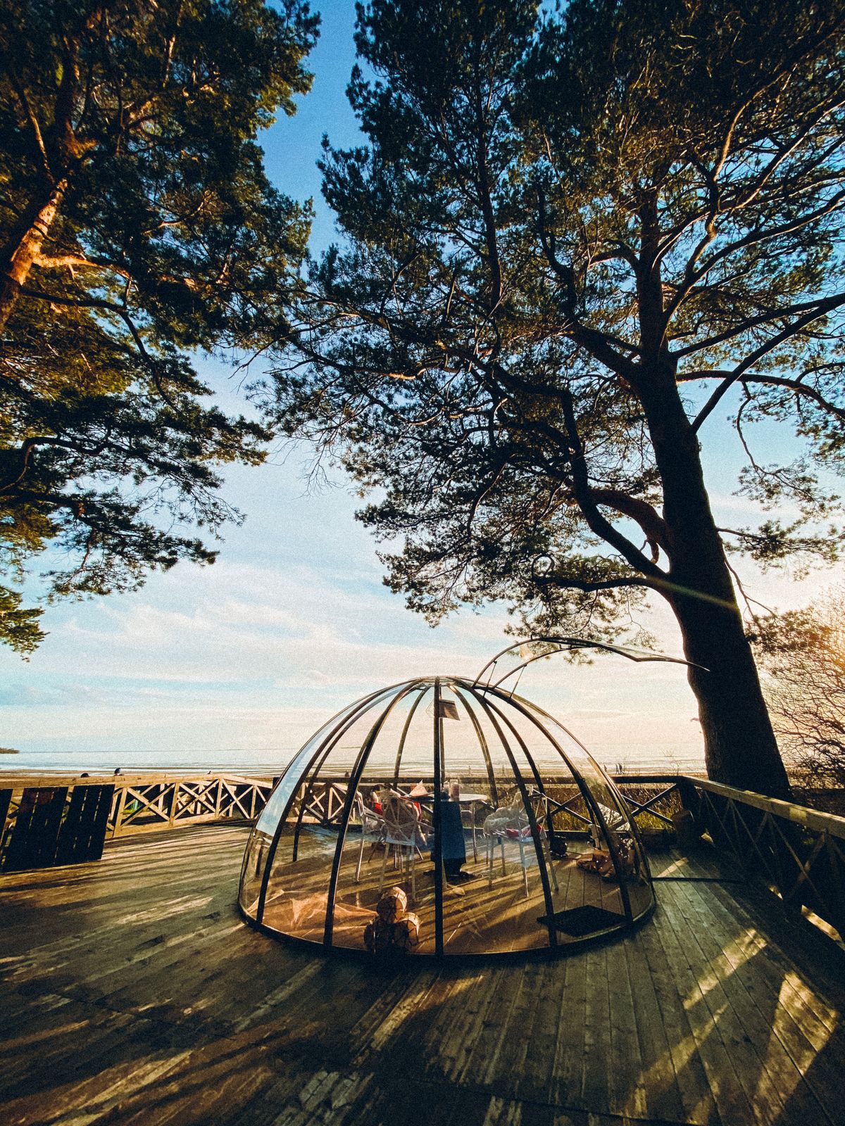 A picturesque glass dome sitting on a wooden terrace surrounded by tall trees and overlooking a peaceful lake at sunset.